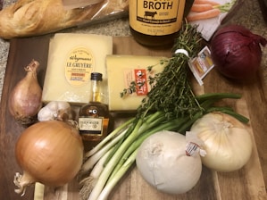 A selection of ingredients is arranged on a wooden cutting board. There is a loaf of crusty French bread, blocks of Gruyère cheese, a bottle of cognac, and a large bottle of beef broth. Surrounding these items are various vegetables, including garlic, shallots, green onions, and red and yellow onions. Fresh thyme sprigs are also present, tied with a string.