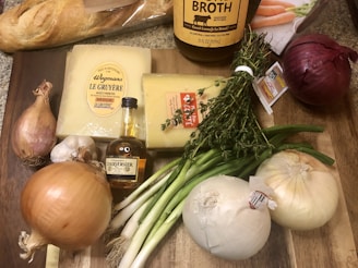 A selection of ingredients is arranged on a wooden cutting board. There is a loaf of crusty French bread, blocks of Gruyère cheese, a bottle of cognac, and a large bottle of beef broth. Surrounding these items are various vegetables, including garlic, shallots, green onions, and red and yellow onions. Fresh thyme sprigs are also present, tied with a string.