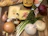 A selection of ingredients is arranged on a wooden cutting board. There is a loaf of crusty French bread, blocks of Gruyère cheese, a bottle of cognac, and a large bottle of beef broth. Surrounding these items are various vegetables, including garlic, shallots, green onions, and red and yellow onions. Fresh thyme sprigs are also present, tied with a string.