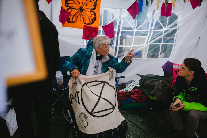 An elderly person sits in a chair holding a banner with a symbol, engaged in conversation with a younger person seated nearby. Brightly colored flags and banners with symbols and designs hang around the tent interior, contributing to a lively and engaged atmosphere.