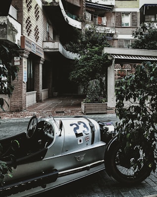A vintage car with number 23 and international flags is parked in an urban courtyard. The surroundings include a multi-story building with exposed brickwork, some vegetation in planters, and a storefront with a sign that reads 'Inmobiliaria Real Estate.' There are balconies with clothes hanging and a generally well-worn but charming urban environment.