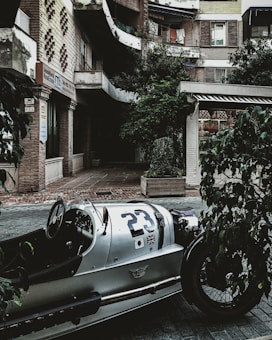 A vintage car with number 23 and international flags is parked in an urban courtyard. The surroundings include a multi-story building with exposed brickwork, some vegetation in planters, and a storefront with a sign that reads 'Inmobiliaria Real Estate.' There are balconies with clothes hanging and a generally well-worn but charming urban environment.