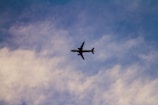A commercial airplane is flying through the sky, silhouetted against a backdrop of wispy clouds and a gradient of blue and white.