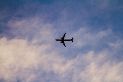 A commercial airplane is flying through the sky, silhouetted against a backdrop of wispy clouds and a gradient of blue and white.