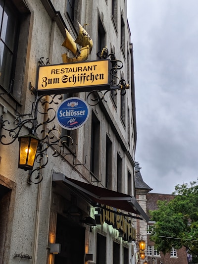A restaurant sign with intricate wrought iron detailing and a golden ship emblem. Below it, a blue circular sign with the word 'Schlösser Alt' hangs. The building is made of stone with large windows and lanterns emitting a warm glow. The sky is overcast, and greenery is visible in the background.