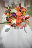A colorful floral bouquet composed of vibrant orange roses, yellow billy balls, white freesias, and purple carnations. The arrangement is accented with green leaves and dark decorative spirals, held by a person wearing a white dress.