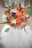 A colorful floral bouquet composed of vibrant orange roses, yellow billy balls, white freesias, and purple carnations. The arrangement is accented with green leaves and dark decorative spirals, held by a person wearing a white dress.