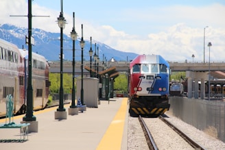 A train with a red, white, and blue front is arriving at a train station with a mountainous backdrop. Several lampposts line the platform, which is mostly empty except for some seating areas. The sky is partly cloudy, and there is a bridge in the background.