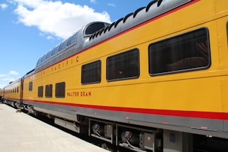 A vibrant yellow train car with the name 'Walter Dean' emblazoned in red letters is parked on a clear day. The train features a double-decker section with large windows and a sleek, rounded roof. The words 'Union Pacific' appear prominently along the upper side, and the train is situated on smooth, white concrete. A bright blue sky with scattered clouds serves as the backdrop.
