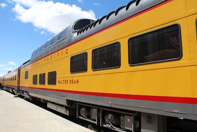 A vibrant yellow train car with the name 'Walter Dean' emblazoned in red letters is parked on a clear day. The train features a double-decker section with large windows and a sleek, rounded roof. The words 'Union Pacific' appear prominently along the upper side, and the train is situated on smooth, white concrete. A bright blue sky with scattered clouds serves as the backdrop.