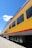 A brightly colored yellow passenger train car with large windows is positioned on railroad tracks. The train has a red stripe along the side and the name 'Walter Dean' is visible. The sky above is clear and blue, with a few scattered clouds.