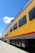 A brightly colored yellow passenger train car with large windows is positioned on railroad tracks. The train has a red stripe along the side and the name 'Walter Dean' is visible. The sky above is clear and blue, with a few scattered clouds.