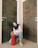 A person wearing a blue hijab and a pink sweatshirt is crouching between two wooden doors, holding a red fire extinguisher. They are wearing blue jeans and white sneakers.