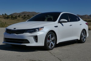 A sleek white sedan parked on a paved road with mountains and clear blue sky in the background. The car features a modern design with sharp lines and well-defined curves, reflecting sunlight off its shiny surface.
