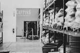 A storage room with shelves lined with bags of coffee beans, wrapped in plastic, labeled with various origins. A sign that reads 'Cupping Laboratory' is visible on the wall. A small forklift is parked in the center, and several plastic buckets are arranged on the floor.
