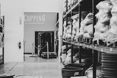 A storage room with shelves lined with bags of coffee beans, wrapped in plastic, labeled with various origins. A sign that reads 'Cupping Laboratory' is visible on the wall. A small forklift is parked in the center, and several plastic buckets are arranged on the floor.