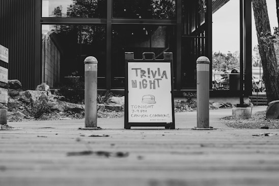 A black and white image featuring a sandwich board sign advertising 'Trivia Night' with details about the event taking place at 'Canyon Commons.' The board is placed on a wooden walkway, flanked by two bollards. In the background, there is a modern building with large glass windows and some foliage.