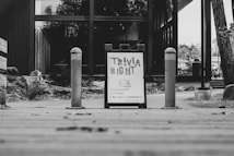 A black and white image featuring a sandwich board sign advertising 'Trivia Night' with details about the event taking place at 'Canyon Commons.' The board is placed on a wooden walkway, flanked by two bollards. In the background, there is a modern building with large glass windows and some foliage.