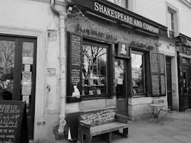 A black and white photograph of a quaint, historic bookstore with wooden-framed windows displaying books. Signage includes 'Shakespeare and Company' and handwritten chalkboard messages. A bench and small round table with a chair are placed outside on the cobblestone street, adding to the cozy European aesthetic.