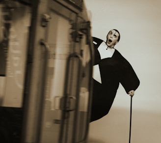 A black and white image of a person in a formal suit with exaggerated expressions and posture. The person holds a cane and appears startled or comical, with a blurred vehicle passing by in the foreground.