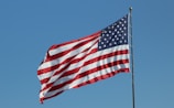An American flag flies against a clear blue sky. The flag features stars and stripes in red, white, and blue, symbolizing the United States.