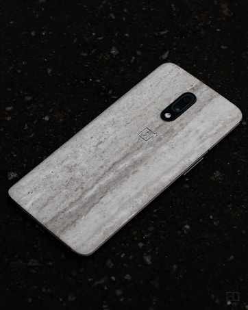 A smartphone with a sleek textured back cover lying on a dark, rough surface. The dual-camera setup is located at the top center. The phone&rsquo;s design features a minimalist aesthetic.