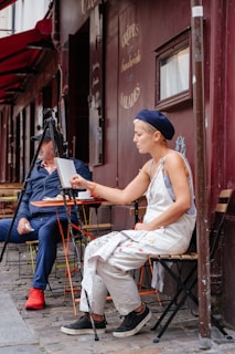 A person is sitting on a chair outdoors, intently painting on a canvas set up on an easel. They are wearing a beret and overalls, which are splattered with paint. Another person sits nearby, dressed in casual clothing, appearing to converse or observe. The setting appears to be a street-side café with a sign reading 'Crêpes Sandwiches Salades' on the dark maroon-colored wall behind them.