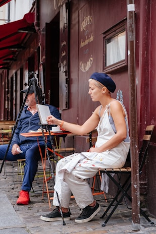 A person is sitting on a chair outdoors, intently painting on a canvas set up on an easel. They are wearing a beret and overalls, which are splattered with paint. Another person sits nearby, dressed in casual clothing, appearing to converse or observe. The setting appears to be a street-side café with a sign reading 'Crêpes Sandwiches Salades' on the dark maroon-colored wall behind them.