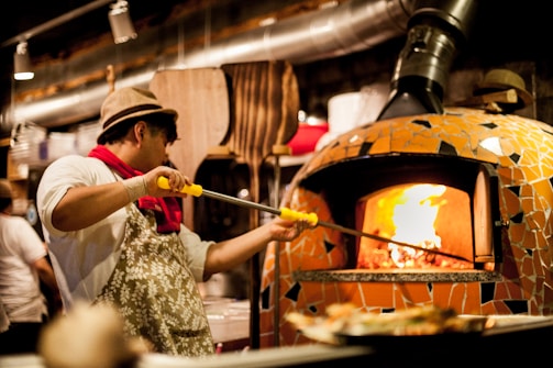 A person wearing a hat and apron is using a long-handled tool to place a pizza into a large, mosaic-tiled wood-fired oven. The oven is glowing with warm light from the fire inside. The background shows various kitchen utensils and another person is seen working.