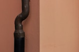A vertical pipe with a curved elbow joint is affixed to a plain, peach-colored wall. The pipe is dark, possibly made of metal, and seems to be secured to the wall with small bolts.