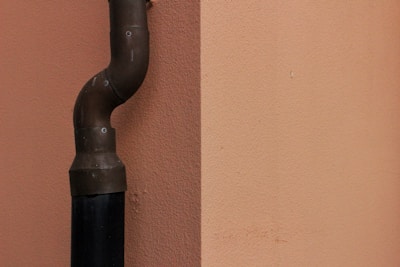 A vertical pipe with a curved elbow joint is affixed to a plain, peach-colored wall. The pipe is dark, possibly made of metal, and seems to be secured to the wall with small bolts.