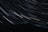 A dark mountainous landscape is illuminated by the long-exposure capture of numerous star trails, creating arcs of light across the night sky.
