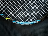 A close-up view of a tennis racket featuring a mesh pattern of white strings against a dark background. The rim of the racket is visible with blue, black, and yellow accents, and the text 'ENGINEERED POWER' is prominently displayed on the frame.