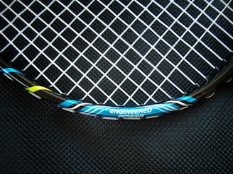 A close-up view of a tennis racket featuring a mesh pattern of white strings against a dark background. The rim of the racket is visible with blue, black, and yellow accents, and the text 'ENGINEERED POWER' is prominently displayed on the frame.