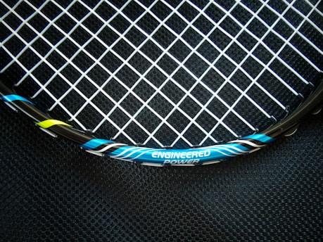 A close-up view of a tennis racket featuring a mesh pattern of white strings against a dark background. The rim of the racket is visible with blue, black, and yellow accents, and the text 'ENGINEERED POWER' is prominently displayed on the frame.
