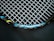 A close-up view of a tennis racket featuring a mesh pattern of white strings against a dark background. The rim of the racket is visible with blue, black, and yellow accents, and the text 'ENGINEERED POWER' is prominently displayed on the frame.