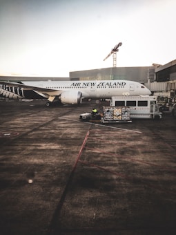 A large passenger airplane from Air New Zealand is parked at an airport terminal. Ground crew and equipment, including a loading cart, surround the aircraft. The terminal building and an industrial crane are visible in the background.