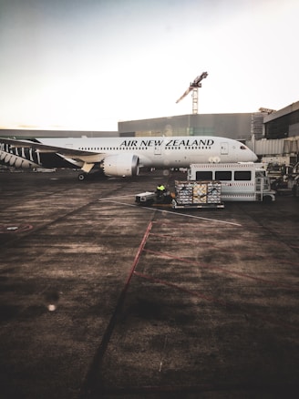 A large passenger airplane from Air New Zealand is parked at an airport terminal. Ground crew and equipment, including a loading cart, surround the aircraft. The terminal building and an industrial crane are visible in the background.