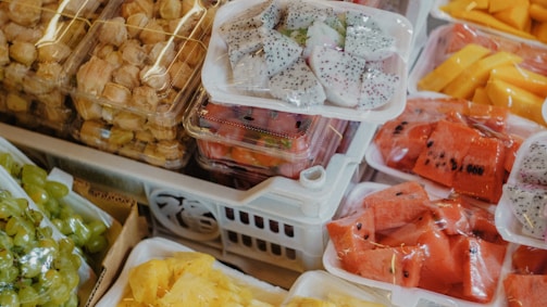 A variety of fresh fruits displayed in plastic trays at a market. The assortment includes bright red watermelon slices, white dragon fruit with black seeds, peeled yellow mango slices, green grapes, and small packaged fruit.