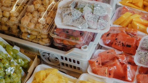 A variety of fresh fruits displayed in plastic trays at a market. The assortment includes bright red watermelon slices, white dragon fruit with black seeds, peeled yellow mango slices, green grapes, and small packaged fruit.
