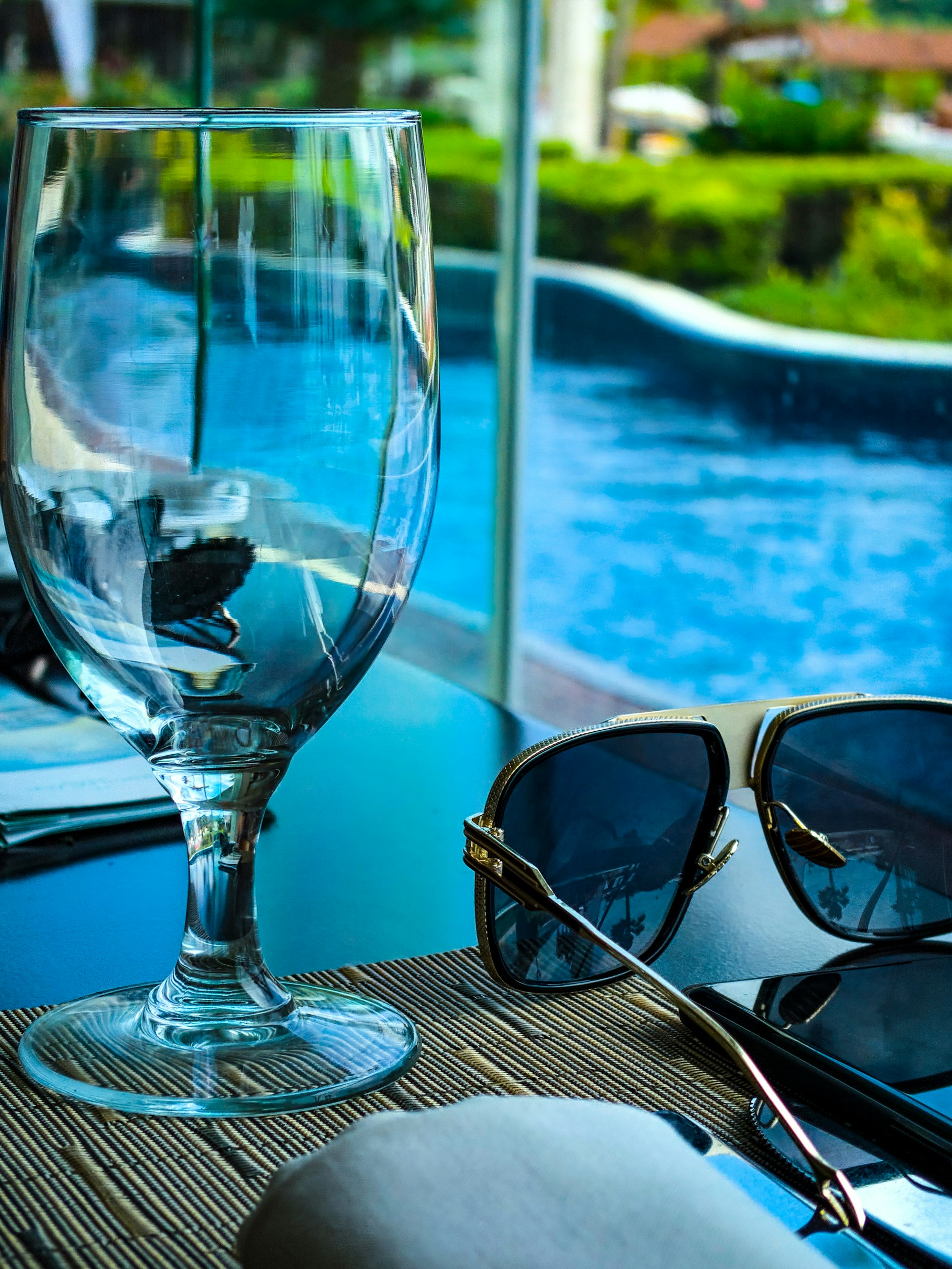 High fashion sunglasses, goblet and phone resting on a restaurant table. In front of a pool, grass and Palm trees. 