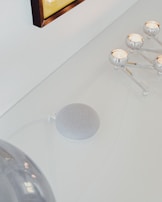 A minimalistic setup on a white surface featuring a smart speaker along with a metallic candle holder with lit candles. The scene includes a partial view of a metallic or glass dome object and a framed picture or artwork on the wall.