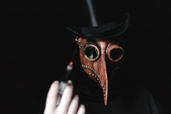 A person wearing a plague doctor mask with a long beak and goggles, dressed in a dark outfit and a black top hat. The mask is adorned with metallic studs, and a hand holding a syringe is visible in the foreground.