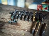 A collection of fishing hooks with metallic components are arranged on a wooden surface. The hooks are attached to metal pieces that resemble bullet casings, with some tips painted in bright colors.