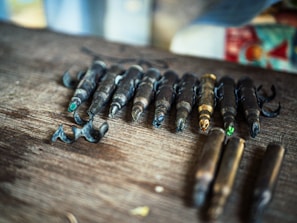 A collection of fishing hooks with metallic components are arranged on a wooden surface. The hooks are attached to metal pieces that resemble bullet casings, with some tips painted in bright colors.