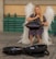 A woman dressed in a floral dress and angel wings is playing a handpan in an outdoor setting. She is seated on a small stool with her eyes closed, seemingly immersed in the music. In front of her are two open cases, one with a 'Donation' sign and the other with a 'CD' sign, indicating she is performing for tips.