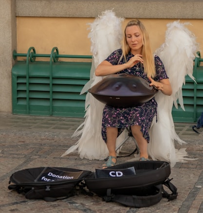 A woman dressed in a floral dress and angel wings is playing a handpan in an outdoor setting. She is seated on a small stool with her eyes closed, seemingly immersed in the music. In front of her are two open cases, one with a 'Donation' sign and the other with a 'CD' sign, indicating she is performing for tips.