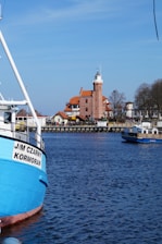 A picturesque coastal scene featuring a serene body of water with vibrant blue hues. A large, colorful boat is moored in the foreground, displaying the name 'J/M Czarny Kormoran' on its side. In the background, a charming brick building with an architectural style reminiscent of a lighthouse adds a touch of historical elegance to the panorama. The sky is clear and blue, enhancing the tranquil atmosphere of the setting.