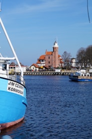 A picturesque coastal scene featuring a serene body of water with vibrant blue hues. A large, colorful boat is moored in the foreground, displaying the name 'J/M Czarny Kormoran' on its side. In the background, a charming brick building with an architectural style reminiscent of a lighthouse adds a touch of historical elegance to the panorama. The sky is clear and blue, enhancing the tranquil atmosphere of the setting.
