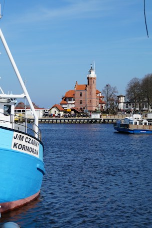 A picturesque coastal scene featuring a serene body of water with vibrant blue hues. A large, colorful boat is moored in the foreground, displaying the name 'J/M Czarny Kormoran' on its side. In the background, a charming brick building with an architectural style reminiscent of a lighthouse adds a touch of historical elegance to the panorama. The sky is clear and blue, enhancing the tranquil atmosphere of the setting.